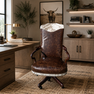A distressed brown leather swivel office chair with a white cowhide backrest and nailhead trim, positioned in a styled home office environment with a desk, wood cabinets, and a rug.