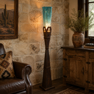 A tall floor lamp with a dark metal base and carved cross design and a textured turquoise-blue glass shade, lit in a rustic living room with a stone wall, leather chair, and wooden cabinet.