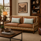 Paddy Southwestern leather loveseat in a warm rustic living room with smooth painted walls, neutral woven rug, wood shelving, desert landscape artwork, and natural sunlight. The loveseat features rich brown leather cushions, geometric patterned upholstery, nailhead trim, and wood framing for a cozy western-inspired look.