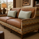 Close-up angled view of a Paddy Southwestern leather loveseat with rich brown leather cushions, geometric patterned fabric upholstery, nailhead trim, and dark wood framing. Styled in a warm rustic living room with soft natural light, neutral woven rug, and desert-inspired decor.