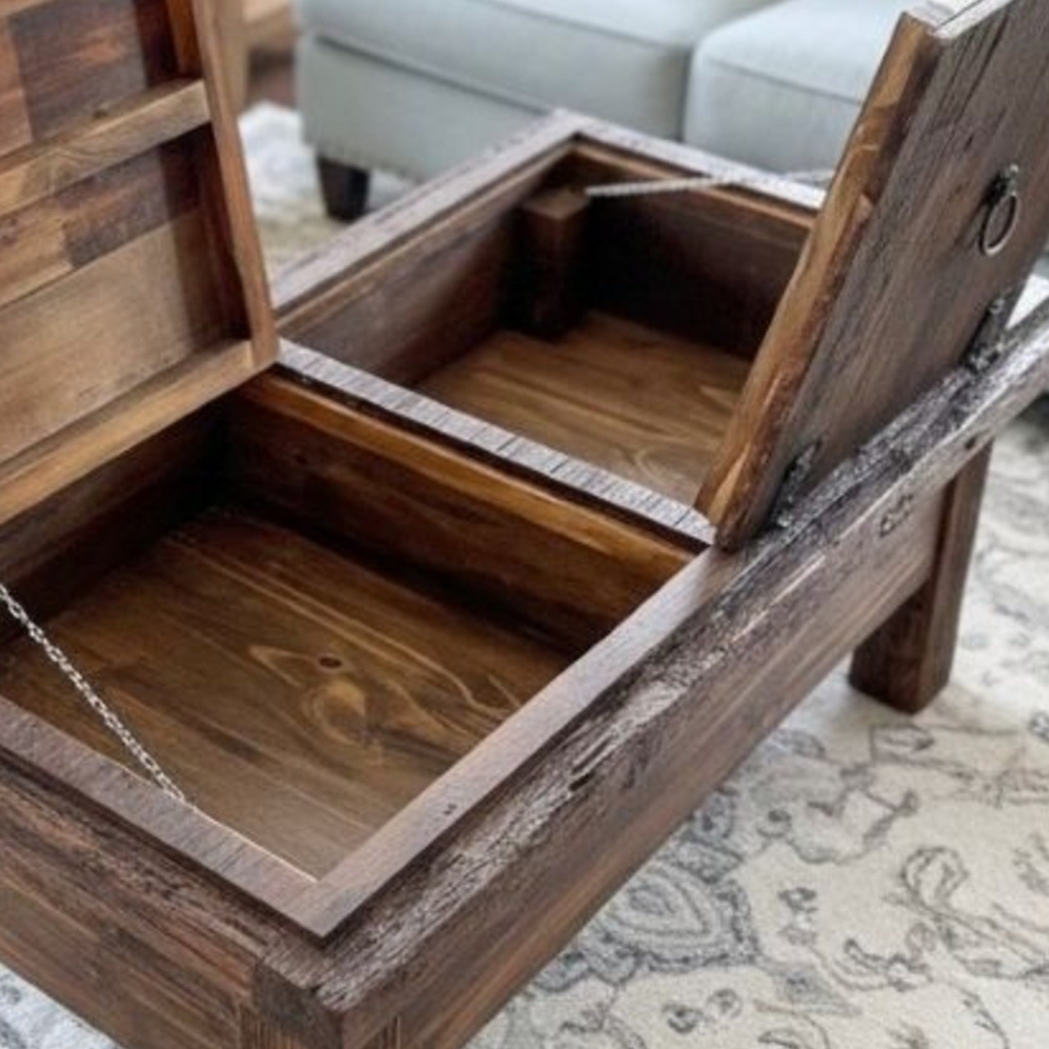 A rustic dark brown wooden storage coffee table with its lid open, showing a chain-latched interior, placed on a patterned rug in a living room setting.