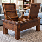 A rustic dark brown wooden storage coffee table with two hinged lids open, revealing a spacious interior compartment. The table is positioned on a patterned area rug in a cozy living room setting with a light blue sofa, a side table, and a bookshelf in the background.