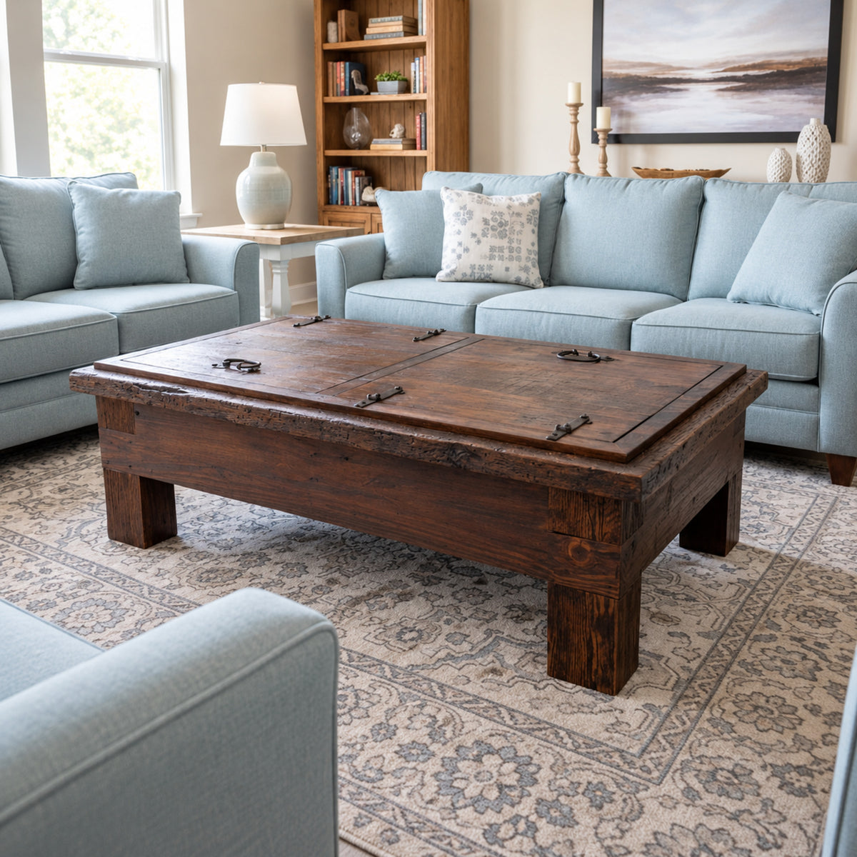 A rustic dark brown wooden coffee table with a hinged lid in a styled living room with light blue sofas and a patterned rug.