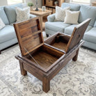 A rustic dark brown wooden storage ottoman with its lid open, revealing a hidden compartment, set on a patterned area rug in a living room with light blue sofas.