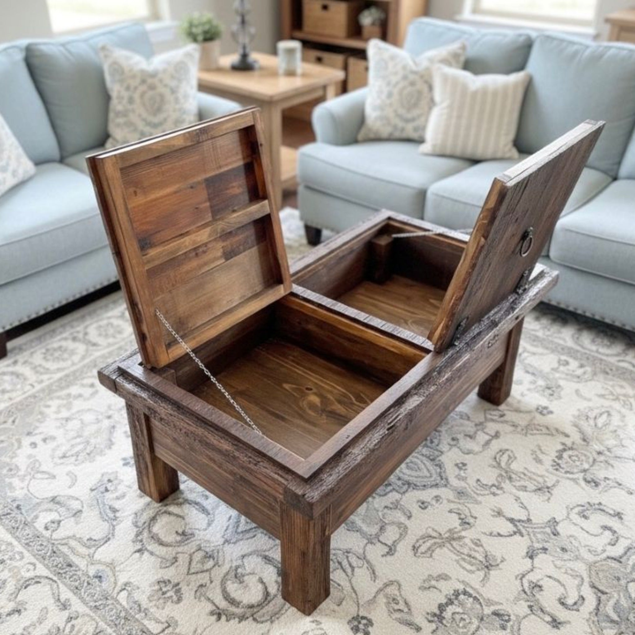 A rustic dark brown wooden storage ottoman with its lid open, revealing a hidden compartment, set on a patterned area rug in a living room with light blue sofas.