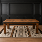 A rustic rectangular wooden coffee table with metal rivet detailing on the apron, shown on a patterned area rug in front of a black paneled wall.