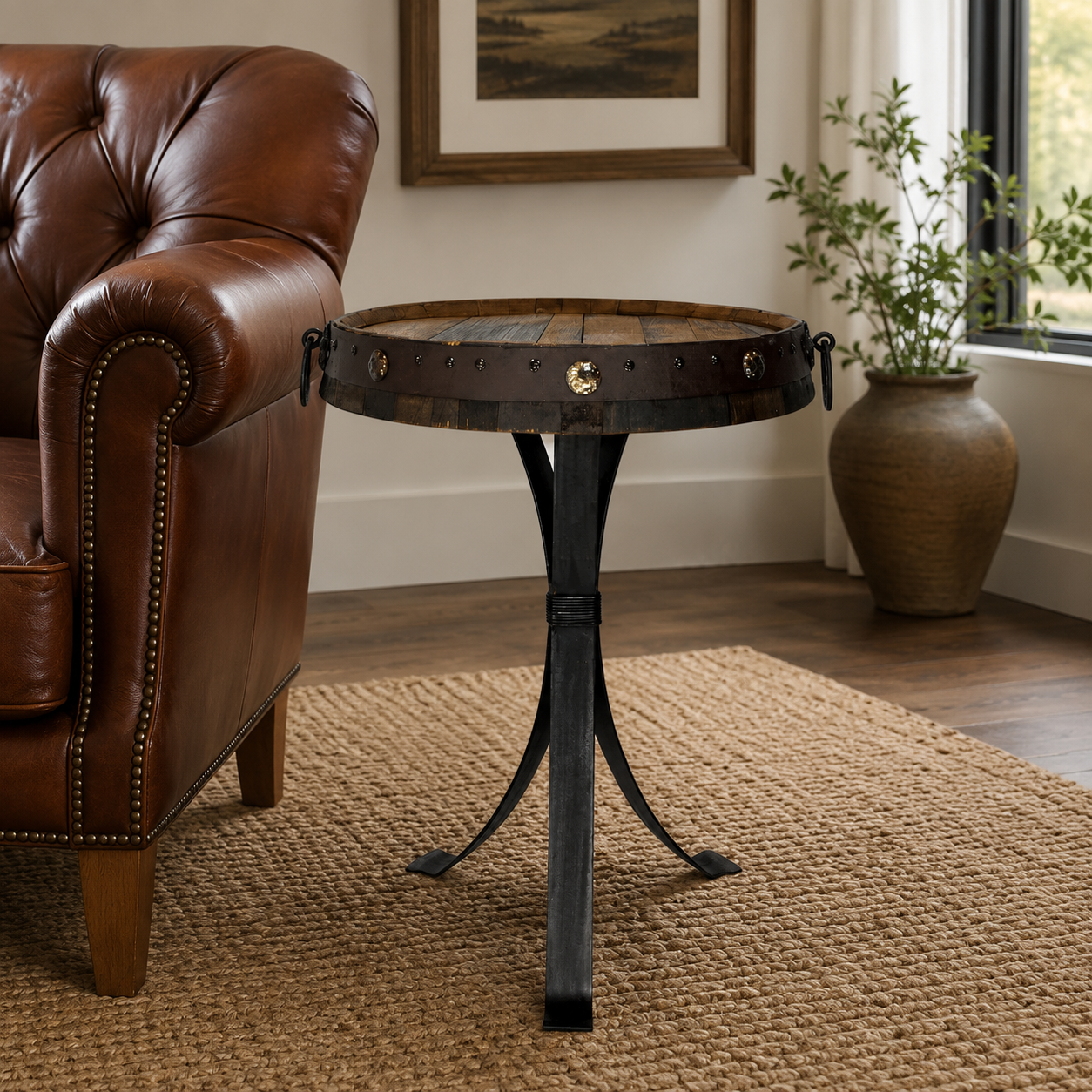 A round rustic end table with a dark wood top and black metal tripod base, styled in a living room next to a brown leather armchair on a woven rug.
