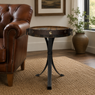 A round rustic end table with a dark wood top and black metal tripod base, styled in a living room next to a brown leather armchair on a woven rug.