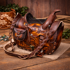 Hand-tooled leather Western saddle purse with braided shoulder strap, embossed horse design, brass latch closure, and rich brown genuine leather, styled on a rustic wooden surface.