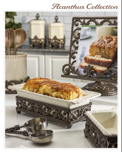 An ornate dark metal stand holding a white ceramic loaf pan with a golden-brown baked loaf inside, set on a white countertop in a kitchen scene. A matching framed picture of a loaf cake stands behind it, with measuring cups in the foreground and the text 'Acanthus Collection' at the top.