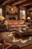 Luxury ranch-inspired living room featuring a tan leather sofa with cowhide accents, set on a cowhide rug in front of a stone fireplace with exposed wood beams, rustic wood tables, and warm western décor 