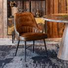 A brown leather dining chair with black iron legs, positioned in a rustic room setting with a wooden wall, a patterned rug, and a stone-topped table.