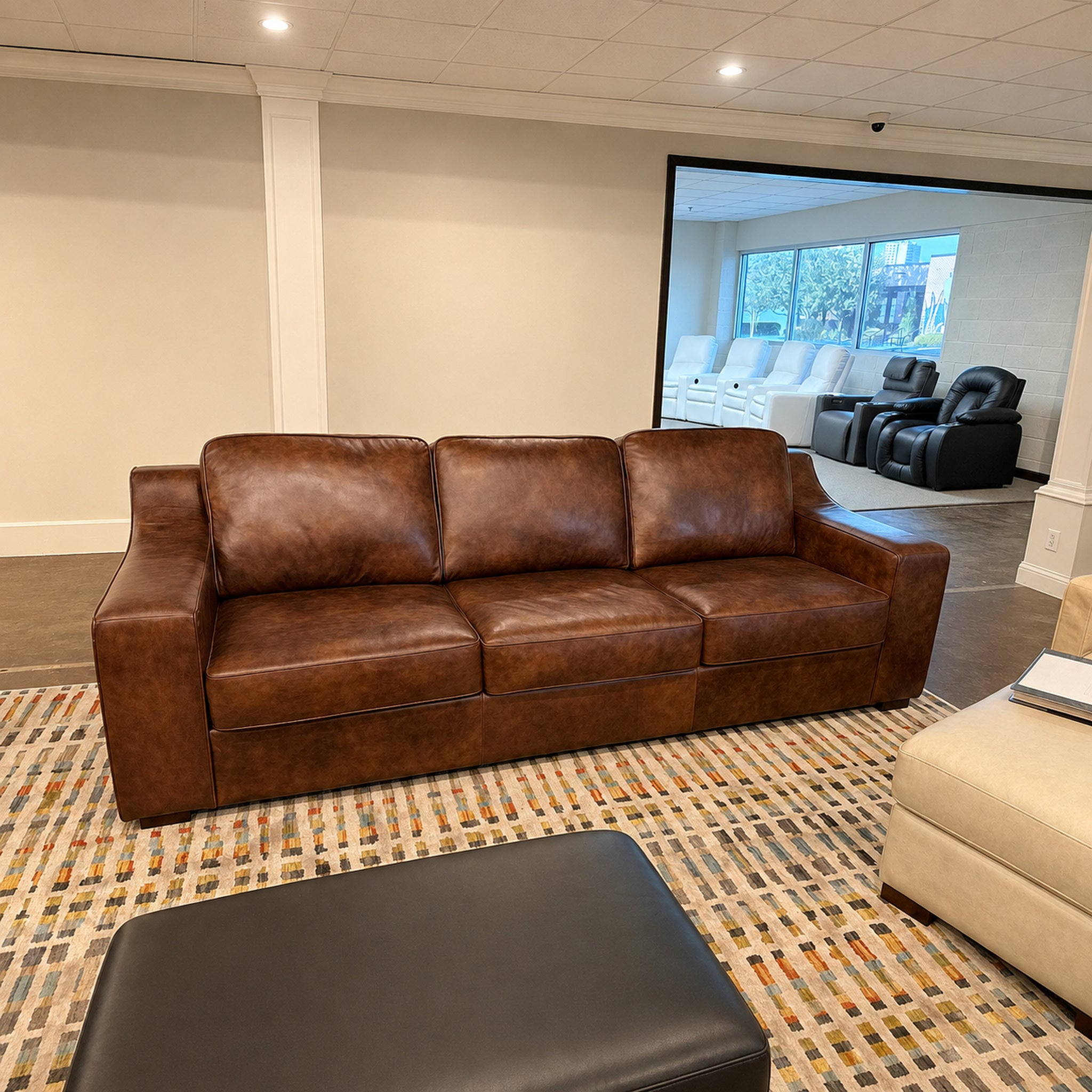A brown leather three-seater sofa in a showroom setting, placed on a patterned rug with a black ottoman in the foreground and a doorway showing other seating in the background.