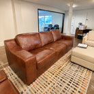 A brown leather three-seater sofa in a modern office or showroom setting, positioned on a patterned geometric area rug next to a cream ottoman.