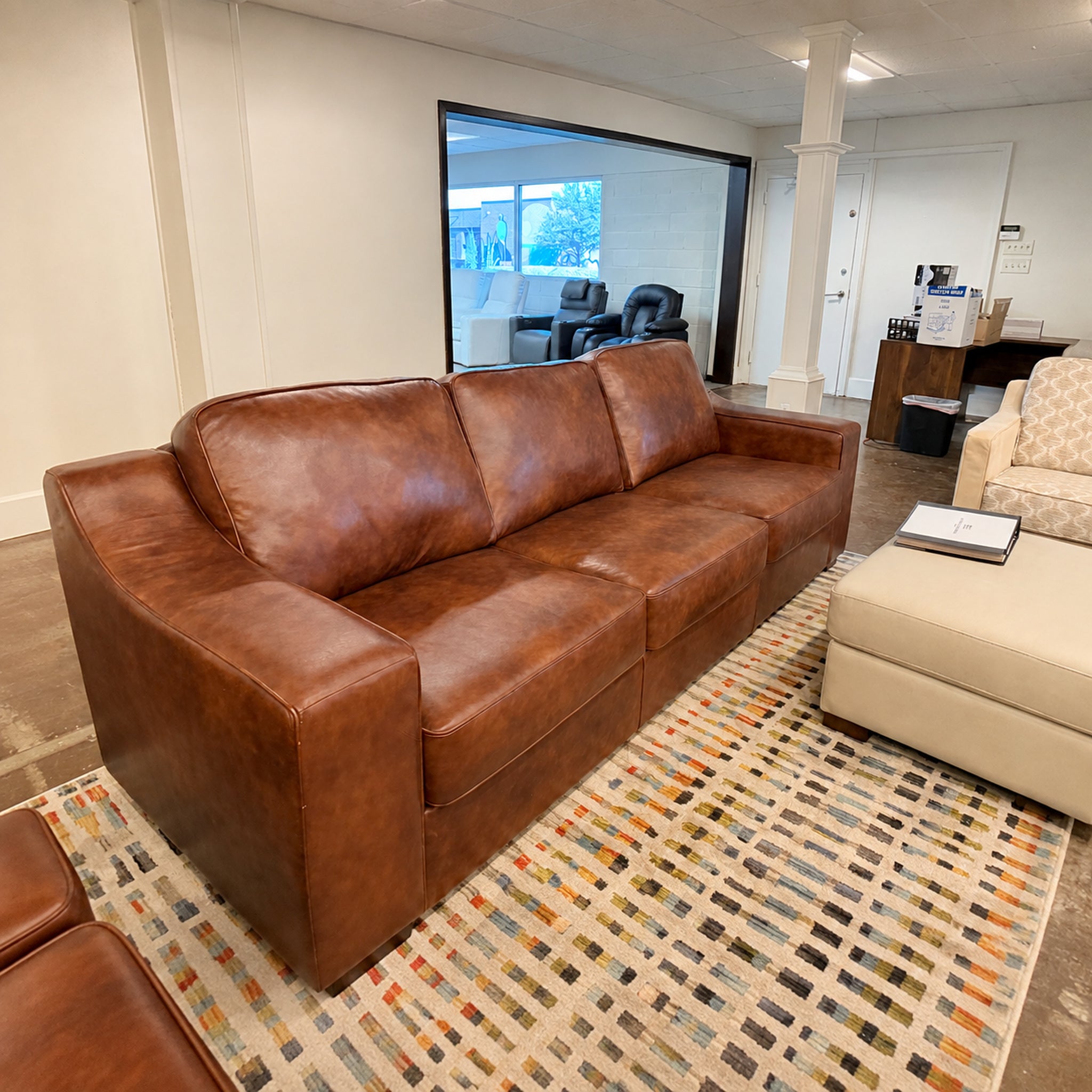 A brown leather three-seater sofa in a modern office or showroom setting, positioned on a patterned geometric area rug next to a cream ottoman.