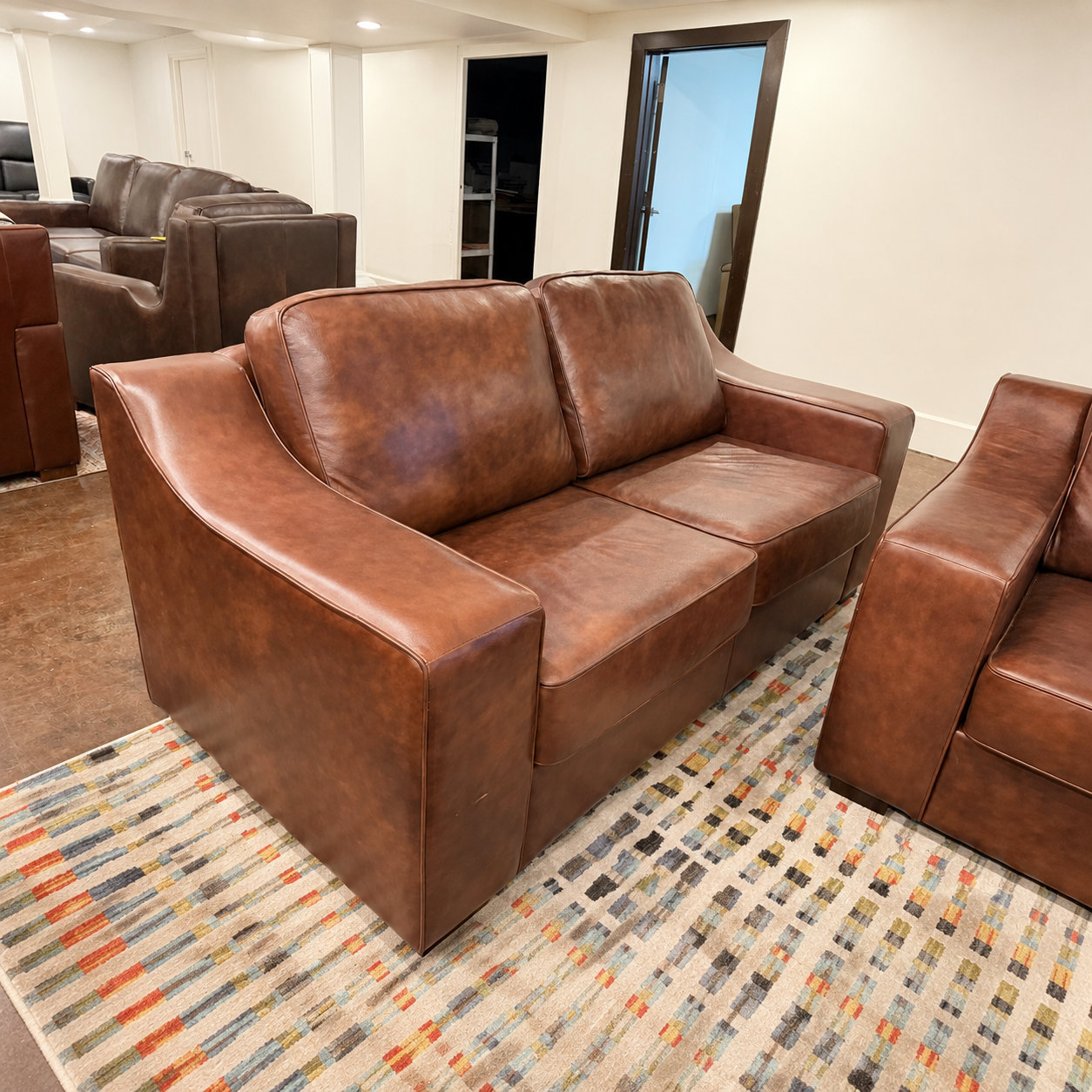 A brown leather loveseat sofa in a home theater or living room setting, positioned on a patterned area rug next to matching brown leather armchairs.