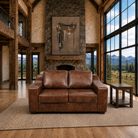 A brown leather loveseat sofa placed in a rustic mountain-view living room with wood floors, large windows, and a stone fireplace.