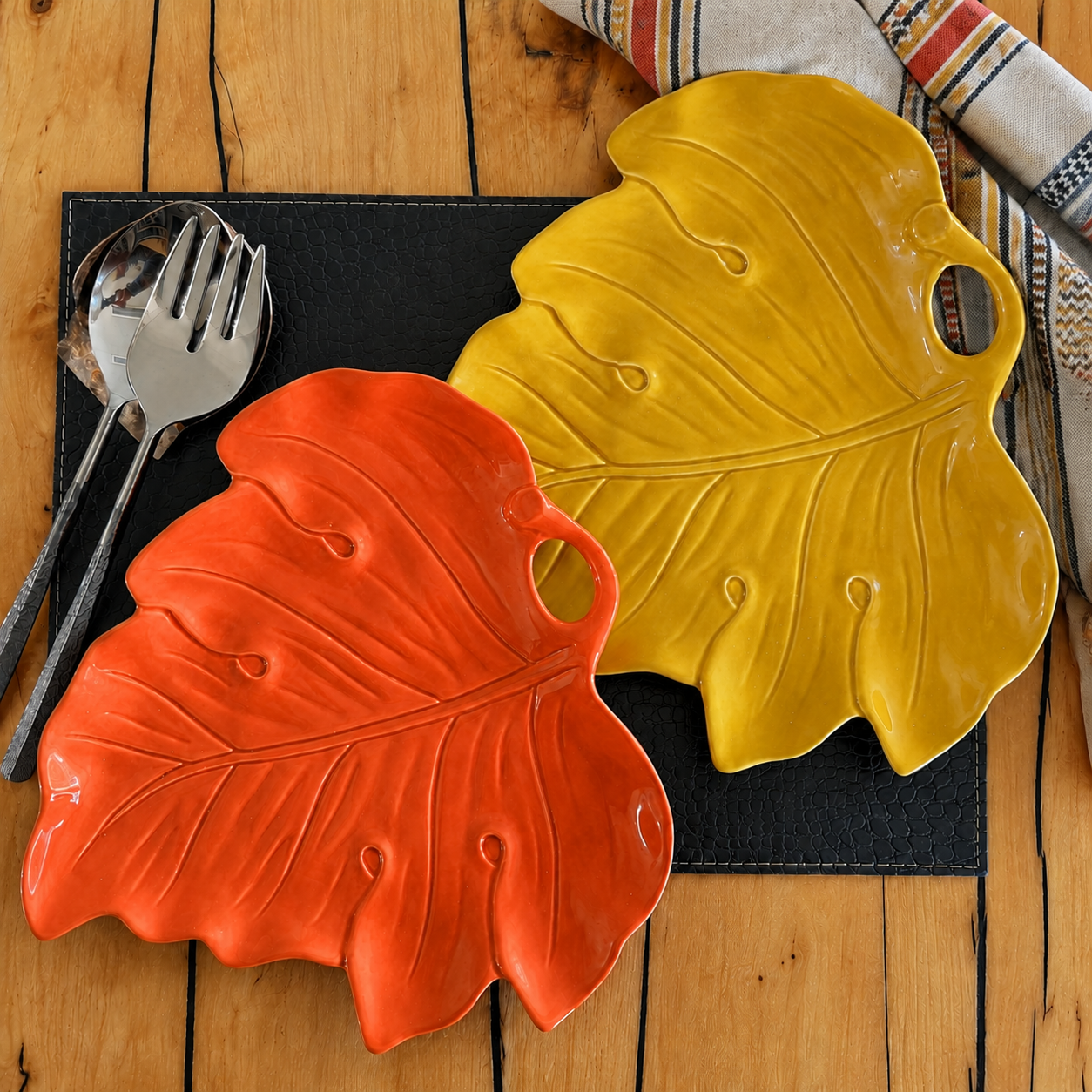 Two ceramic leaf-shaped serving platters in orange and yellow, styled on a black placemat with silver cutlery on a wooden table.