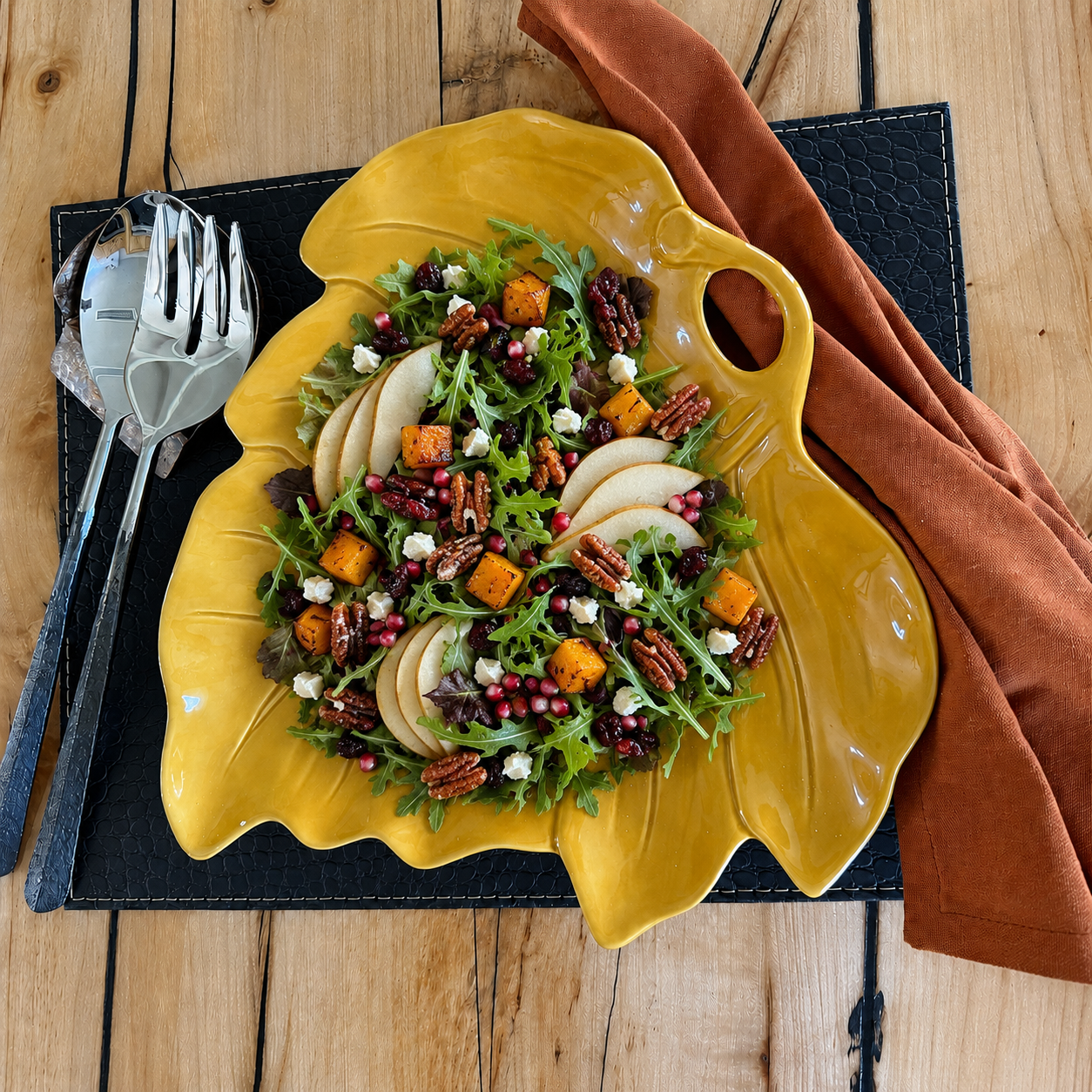 A leaf-shaped yellow ceramic bowl filled with a colorful salad, set on a black placemat with a rust-colored napkin and silver utensils on a wooden table.