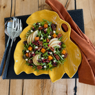 A leaf-shaped yellow ceramic bowl filled with a colorful salad, set on a black placemat with a rust-colored napkin and silver utensils on a wooden table.