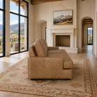 A beige quilted leather sofa in a spacious, modern living room with a stone fireplace, large windows showing a mountain view, and a patterned area rug.