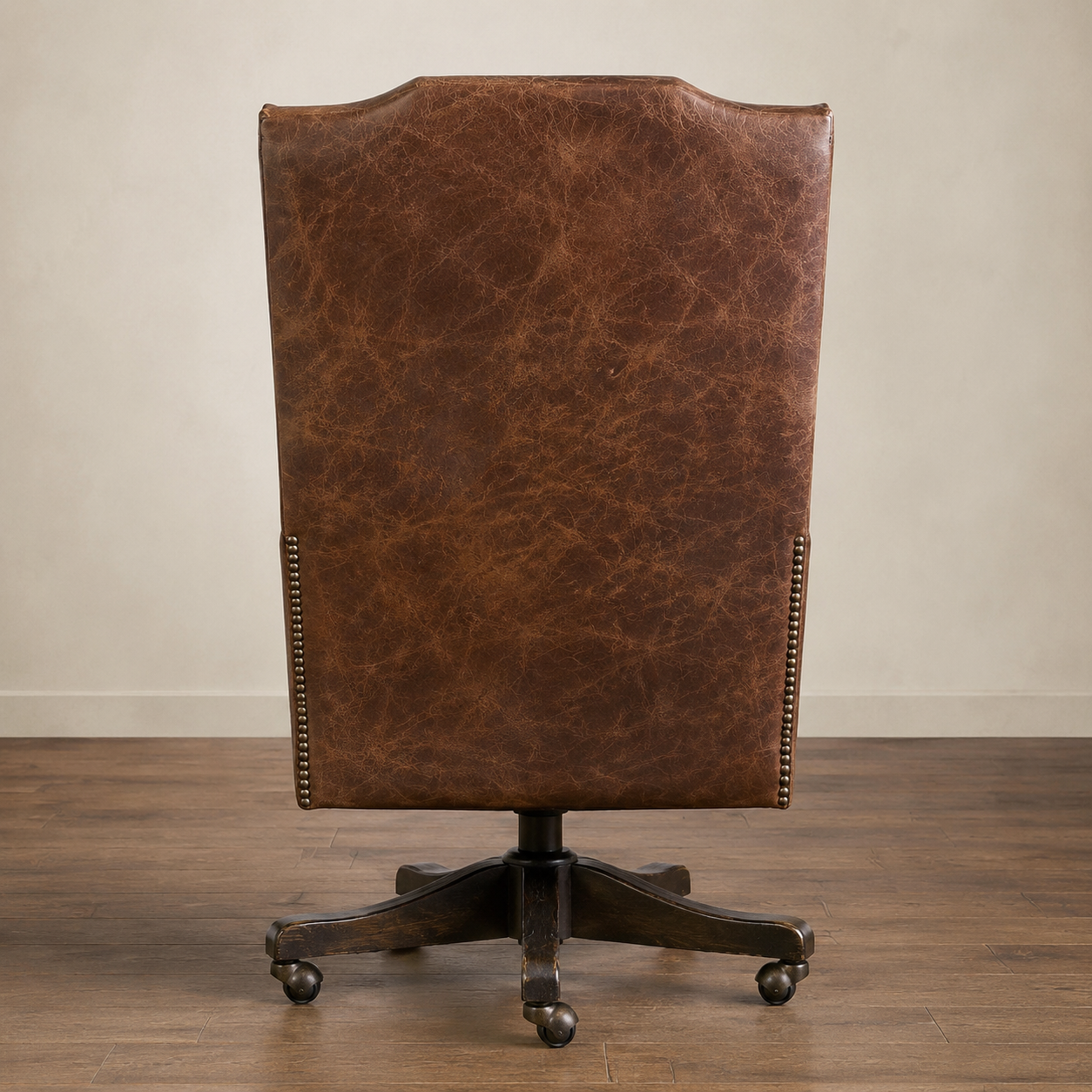 A tall, high-back office chair upholstered in distressed brown leather, featuring nailhead trim along the sides and a dark metal five-star wheeled base, shown against a neutral beige wall and wood floor.