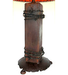 A rustic dark brown wooden table lamp featuring a thick rectangular post wrapped with black barbed wire, a matching dark metal base with a dripping effect, and a red-orange fabric shade partially visible at the top.