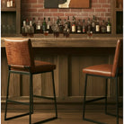 Two bar stools with brown leather seats and textured backs, set in front of a rustic wooden bar counter with liquor bottles and a brick wall background.