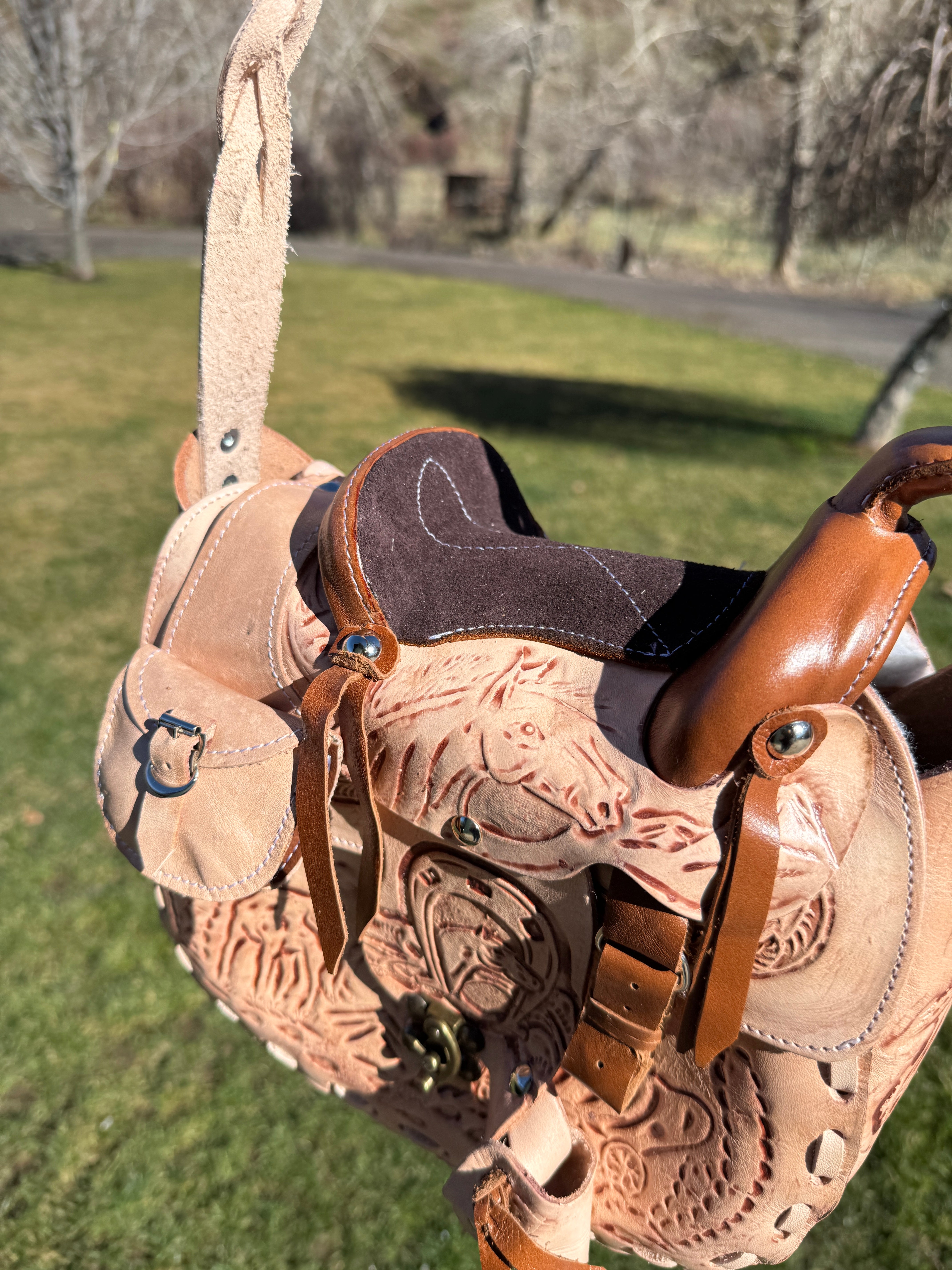 Light beige hand-tooled leather Western saddle purse with embossed horse and horseshoe details, braided strap, and brass latch closure, outdoors