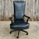A tall black faux-croc leather office chair with dark carved wooden arms and a five-wheel swivel base, set against a rustic wooden fence and concrete floor.