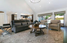 A spacious living room featuring a dark gray leather sofa with patterned throw pillows, two beige armchairs, a round wooden coffee table, and a large patterned area rug. The room has a high vaulted ceiling with recessed lighting and large windows with roller shades.