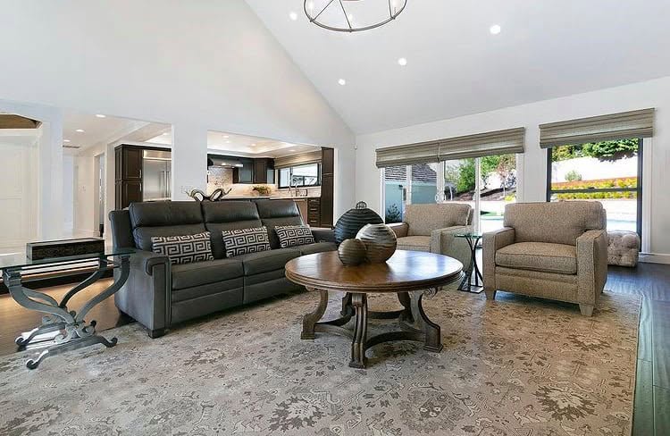 A spacious living room featuring a dark gray leather sofa with patterned throw pillows, two beige armchairs, a round wooden coffee table, and a large patterned area rug. The room has a high vaulted ceiling with recessed lighting and large windows with roller shades.