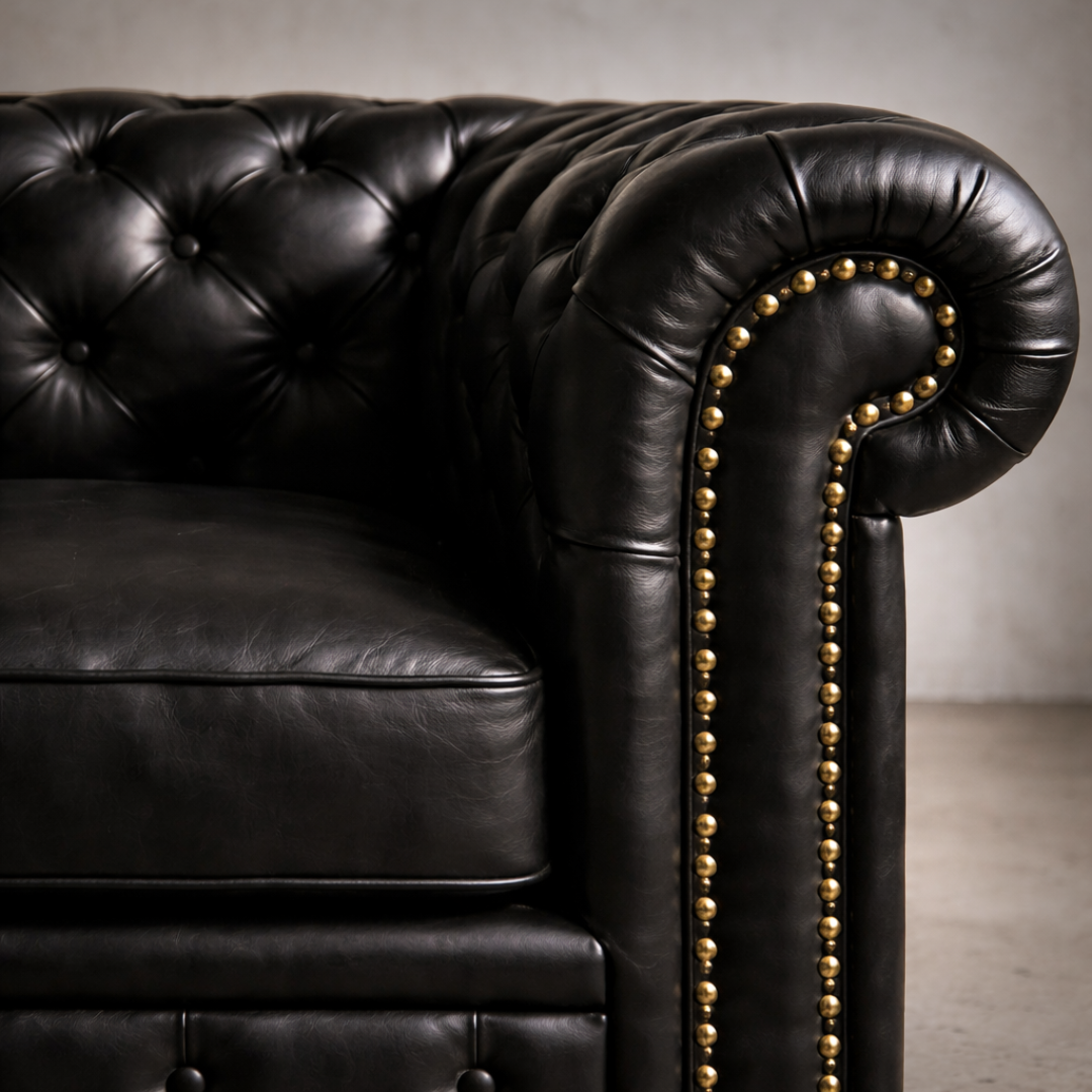 Close-up of a black leather Chesterfield sofa arm with deep button tufting and gold nailhead trim, set against a neutral gray background.