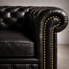 Close-up of a black leather Chesterfield sofa arm with deep button tufting and gold nailhead trim, set against a neutral gray background.