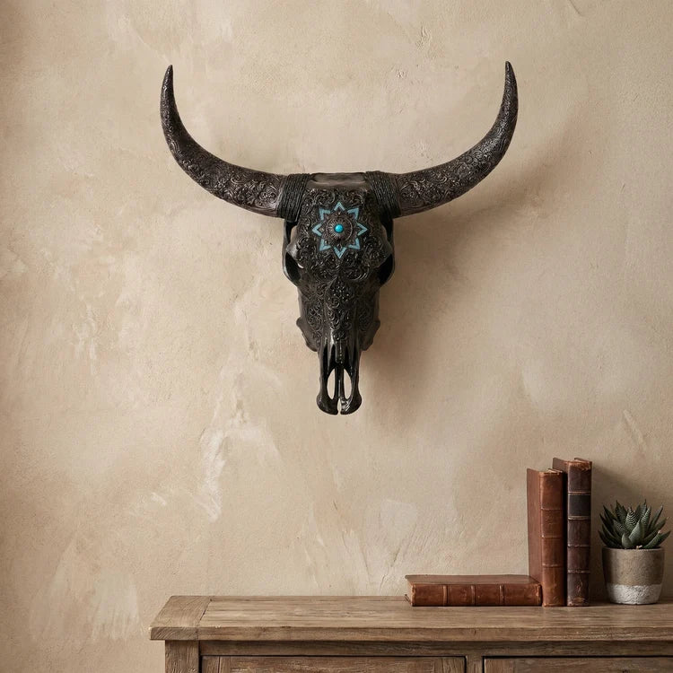 A dark, textured bull skull wall mount with curved horns and a turquoise starburst design on the forehead, displayed on a textured beige wall above a rustic wooden console table with books and a succulent.