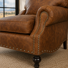 Close-up of a brown leather armchair with rolled arms, nailhead trim, and dark turned legs on a woven rug near a window.