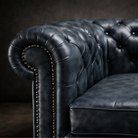 Close-up of a dark blue tufted leather Chesterfield sofa arm with brass nailhead trim against a dark studio background.