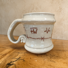 A handmade ceramic mug with a rustic, hand-painted appearance, featuring a light beige/cream glaze and reddish-brown markings of barbed wire and brands, sitting on a wooden surface against a textured beige wall.