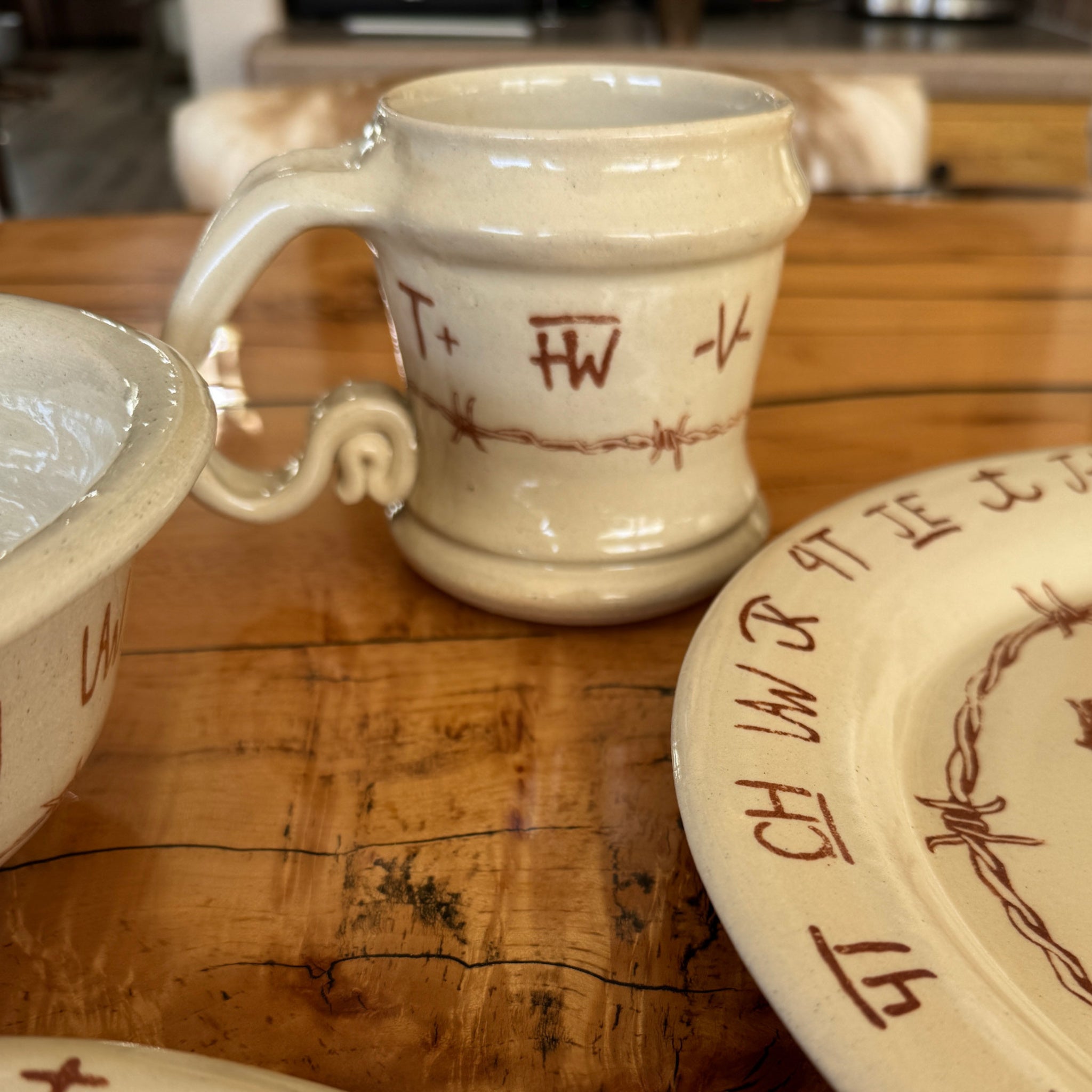 A handmade Blue Mountain Brands ceramic mug with a scroll handle and barbed wire and brands design, sitting on a rustic wooden kitchen island next to matching Blue Mountain Brands dishes.