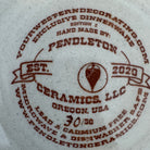 Close-up of a white ceramic dinnerware piece printed with a brown Pendleton Ceramics LLC logo and text, including 'Exclusive Dinnerware' and 'Hand Made By: Pendleton Ceramics, LLC. Exclusive Dinnerware for Your Western Decor