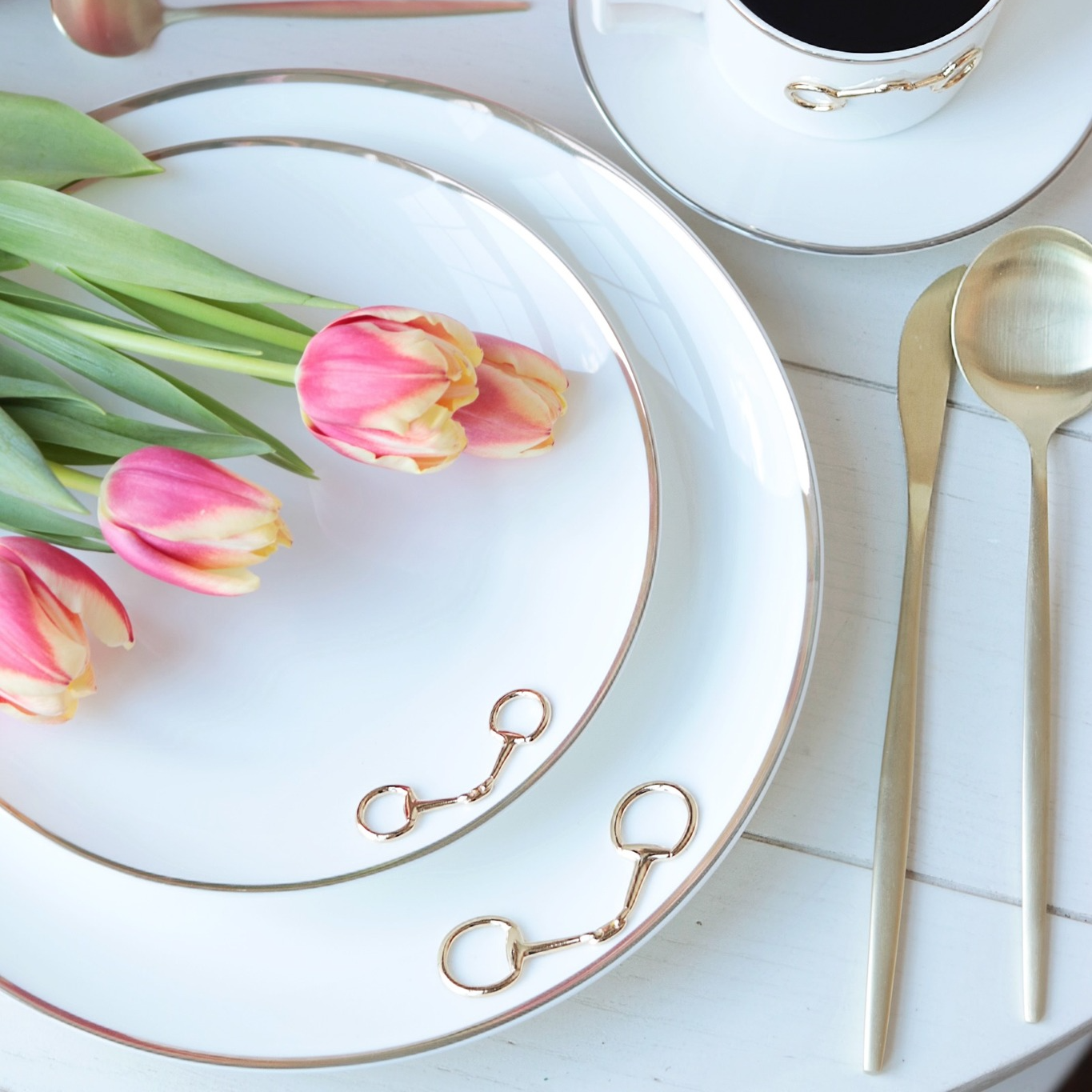 Table setting with pink tulips, gold cutlery, white bone china dishes with gold accents, and decorative elements on a white wood tabletop - Your Western Decor