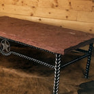 A rustic western-style table with a thick reddish-brown stone top and a black metal frame featuring braided iron legs and a star emblem on the side, set against a wood-paneled wall and cowhide rug.