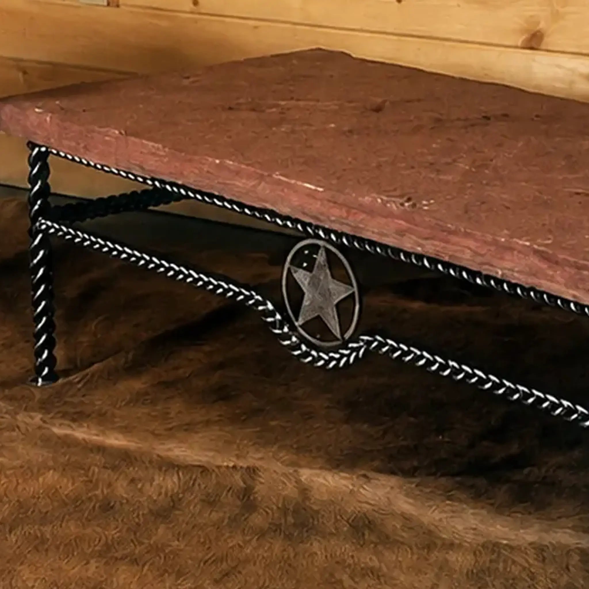 A rustic western-style coffee table with a reddish-brown stone top and a black iron frame featuring braided iron frame and a central western iron star emblem, set against a wood-paneled wall and brown hide rug.