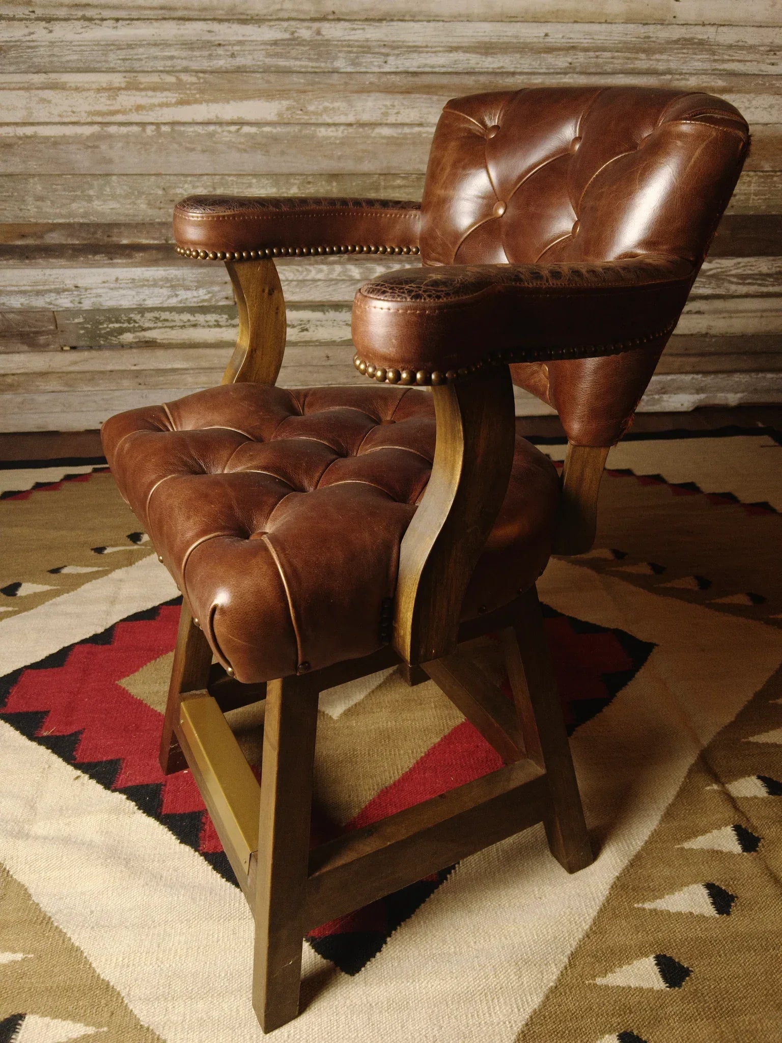 Brown leather bar stool with wooden legs on a patterned rug against a wooden wall - Your Western Decor