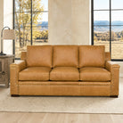 A three-seat leather sofa in a warm tan/caramel color, styled in a bright living room with large windows, a side table, and a lamp.