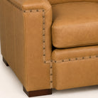 Close-up of a tan leather armchair corner with dark nailhead trim and dark wooden feet on a light studio background.