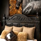A rustic dark wood carved headboard on a bed styled with beige pillows, fluffy tan sherpa pillows, and a cowhide accent pillow, set against a dark wall with a large horse painting above.