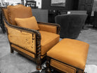 A vintage-style upholstered armchair with a matching ottoman in a warm mustard-yellow fabric, set against a blurred black-and-white showroom background.