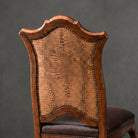 A close-up of the back of a wooden dining chair featuring a tall, shield-shaped backrest with a textured embossed leather panel, dark brown leather seat, and metal nailhead trim, set against a dark gray background.