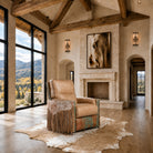 A rustic western-style accent chair with fringe detailing sits on a cowhide rug in a spacious, high-ceilinged room with a stone fireplace and large windows overlooking a mountain landscape.